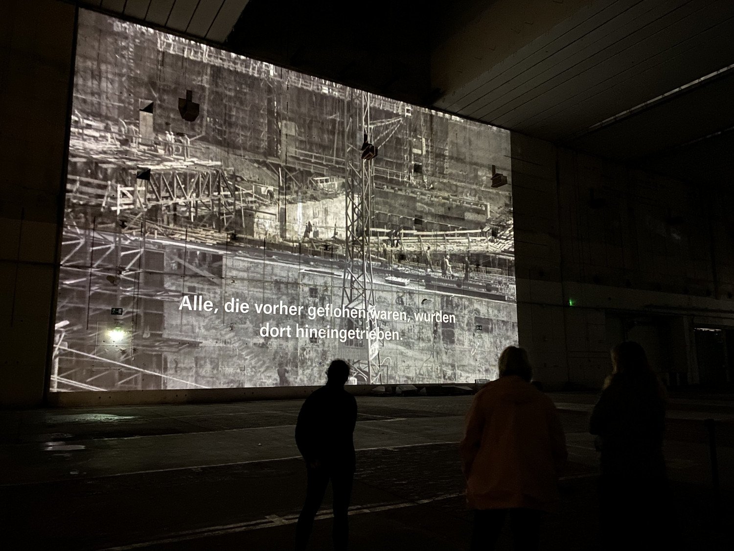 "Alle die vorher geflohen waren, wurden dort hingetrieben" ist im Transkript lesbar. Im Hintergrund sieht man ein Bild der Baustelle in schwarz-weiss. 