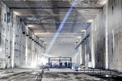 Blick in den Ruinenteil des Bunkers. Mehrere Personen errichten eine Kulisse aus einem Metallgitter. Durch die Bombeneinschläge in der Decke dringt Tageslicht in den dunklen, großen Innenraum. 