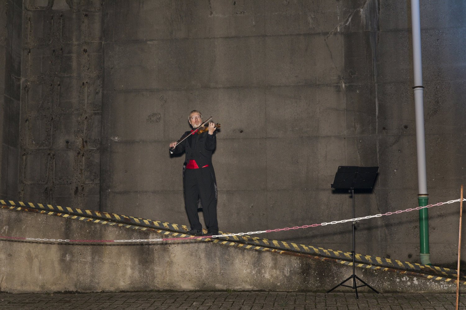 Ein Musiker im Anzug mit einer Geige oder Violine in der Hand spielt vor einer Bunkerwand im Aussenbereich.  