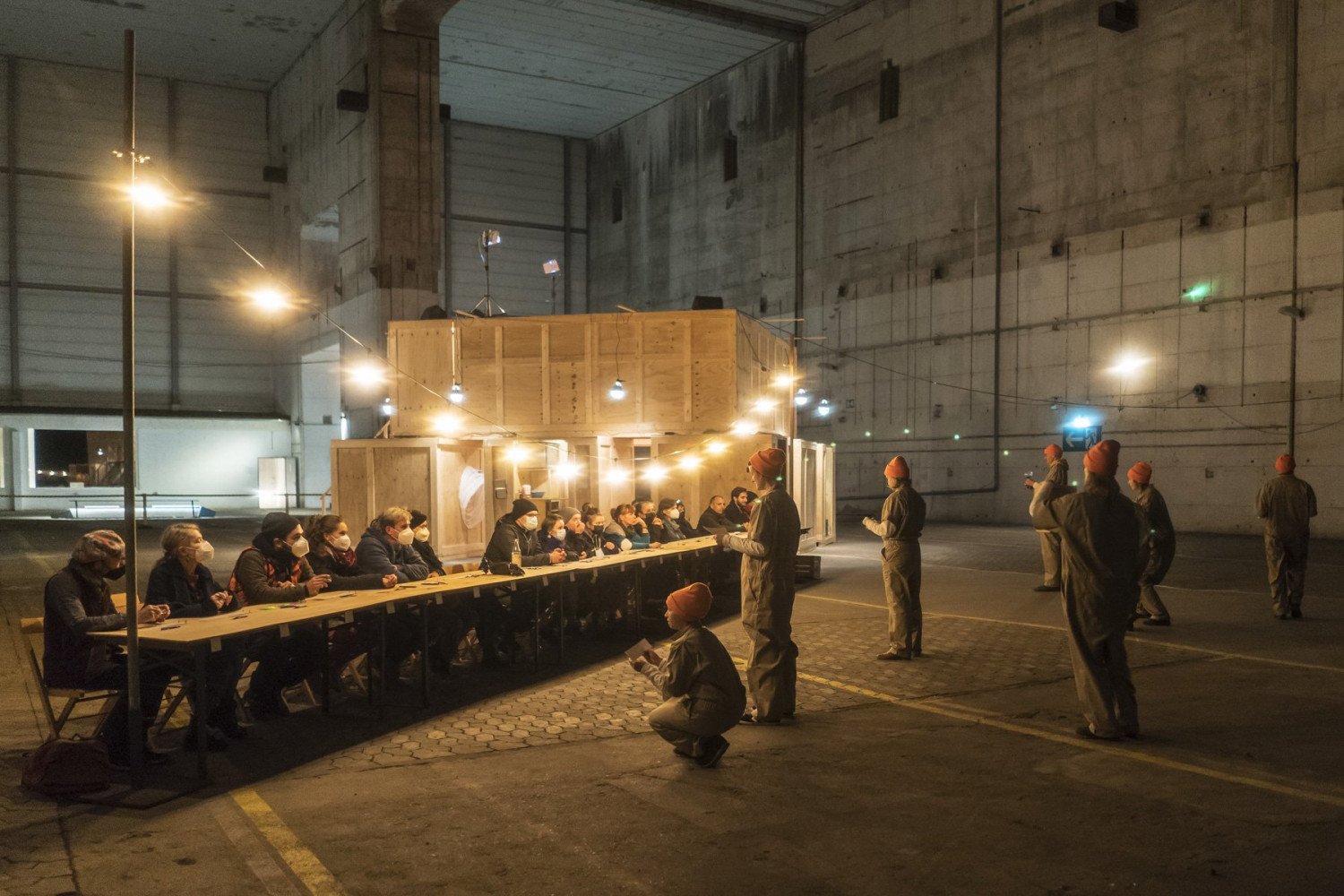 Die Performance-Teilnehmenden, ungefähr 25 Personen, bei ein Probe im Bunkerinnenraum. Einige Personen der Gruppe sitzen an einem sehr langen Tisch, andere stehen in Overalls mit orangsfarbenen Mützen davor.  Eine Person ist in der Hocke. 