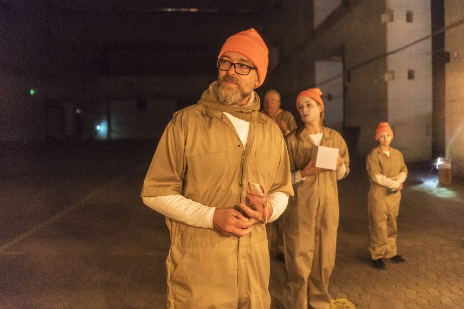 Vier Akteure im Bunkerinnenraum. Sie tragen Overalls und orangefarbene Mützen. Blicken seitlich an der Kamera vorbei. Eine Person hält eine kleine Box in der Hand.  