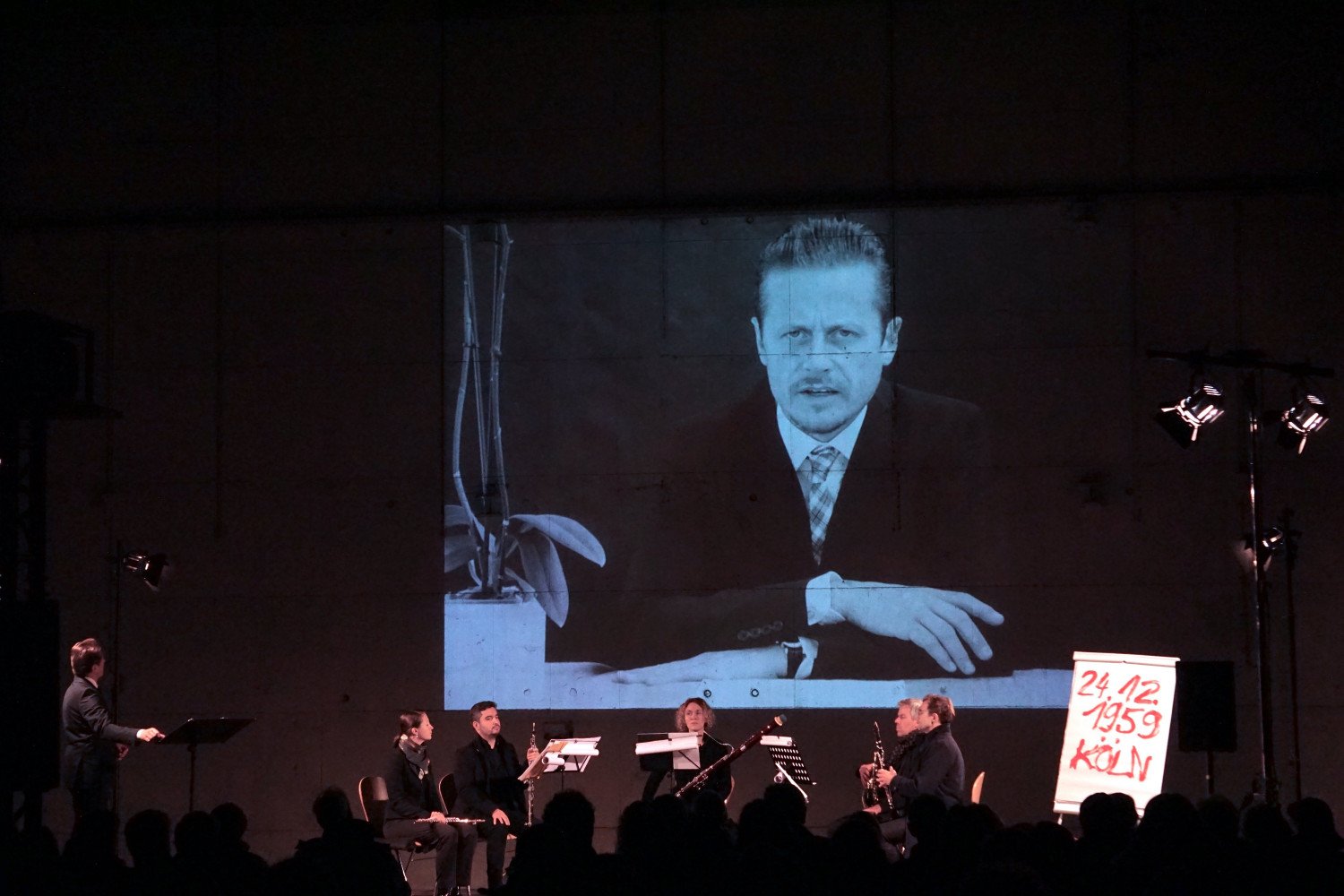 Blick auf das Bühnenbild. Projektion des Schauspielers auf eine im Raum befindliche Leinwand. Davor Musiker:innen sitzend.  