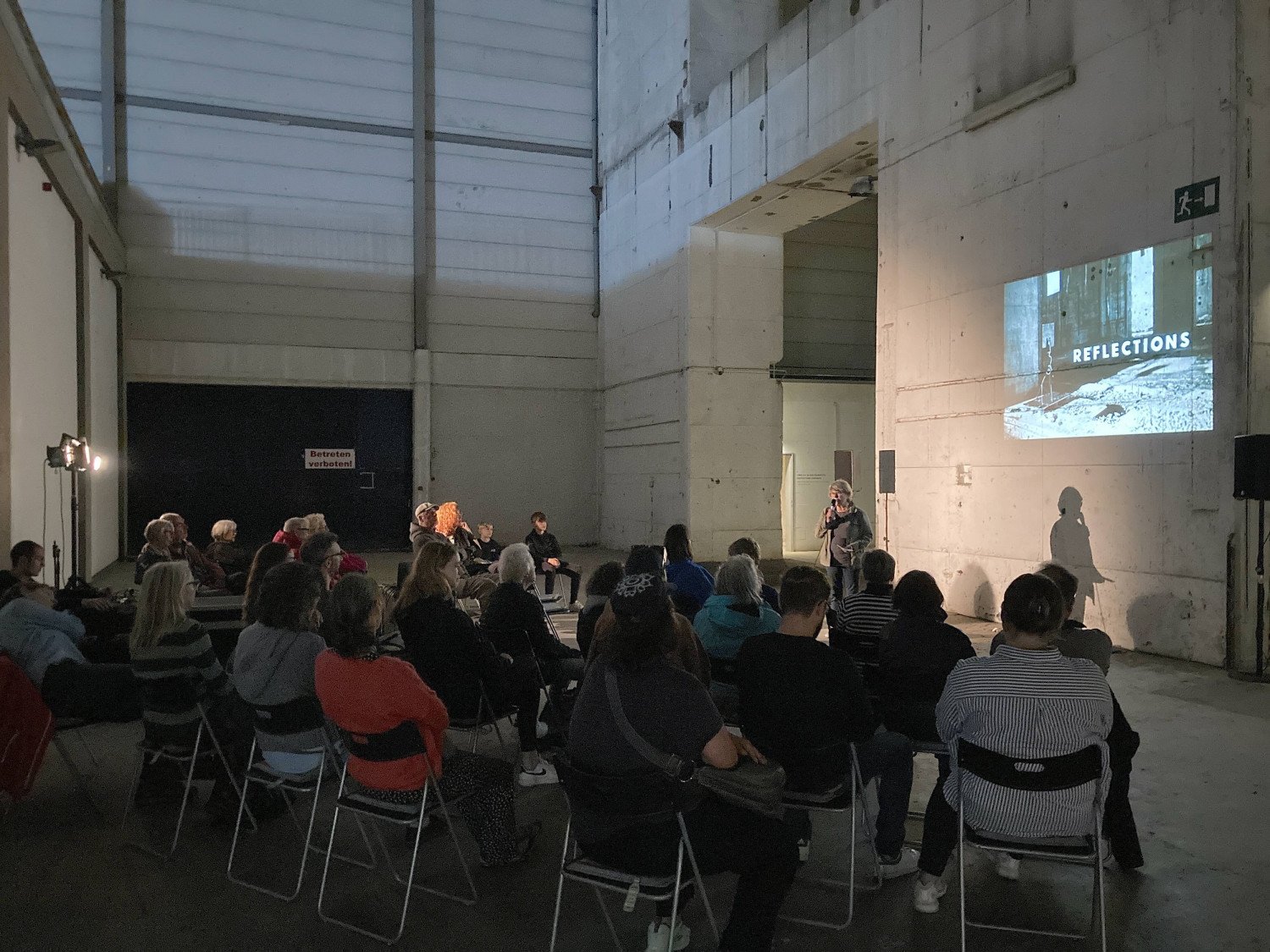 Annemarie Strümpfler steht im Inneren des Bunkers vor einem Publikum. An die Säule hinter ihr wird ein Foto aus dem Ruinenteil des Bunkers mit dem Schriftzug "REFLECTIONS" projiziert 