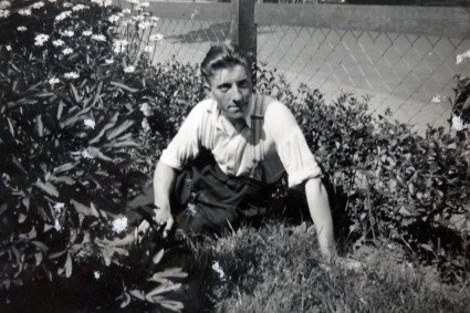 Schwarz-Weiss-Aufnahme, Portrait eines jungen Mannes mit weißen Hemd und hochgekrempelten Ärmeln sitzend auf Gras, umgeben von ein Busch und weiteren blühenden Pflanzen