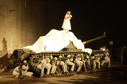 Eine Frau, bekleidet mit einem weißen Brautkleid, steht auf einem Panzer. Im Raupenband des Panzers befinden sich Häftlinge in gestreifter Kleidung, sie tragen eine grüne Girlande. Scheinwerfer erleuchten die Szene.